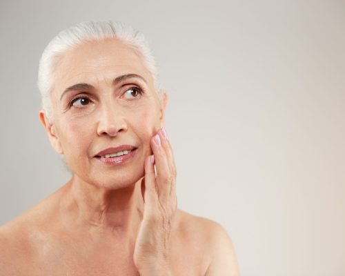 Beauty portrait of an attractive naked elderly woman posing isolated over grey background. Looking aside.